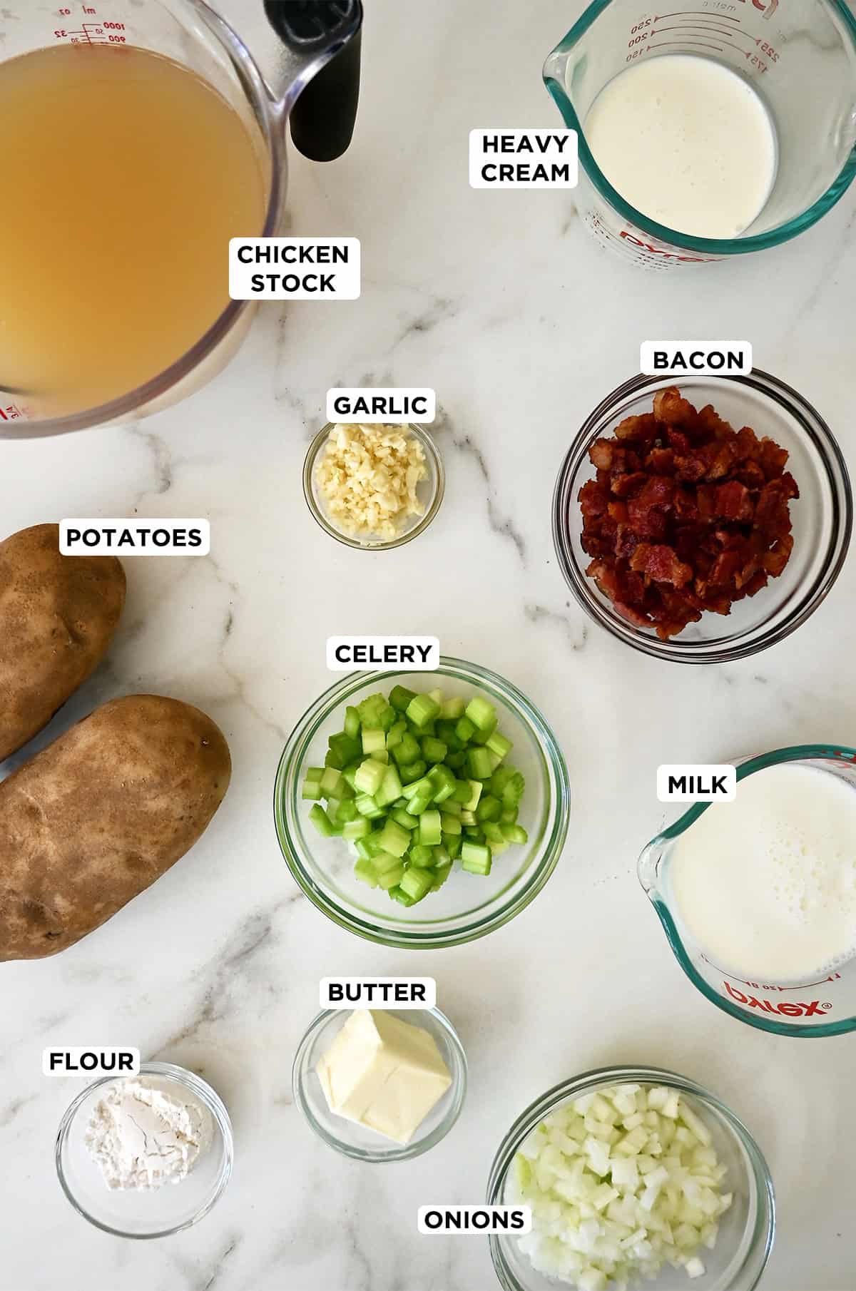 Potato soup ingredients in various sizes of glass bowls, including Russet potatoes, chicken stock, minced garlic, diced celery, bacon, milk, heavy cream, butter and flour.